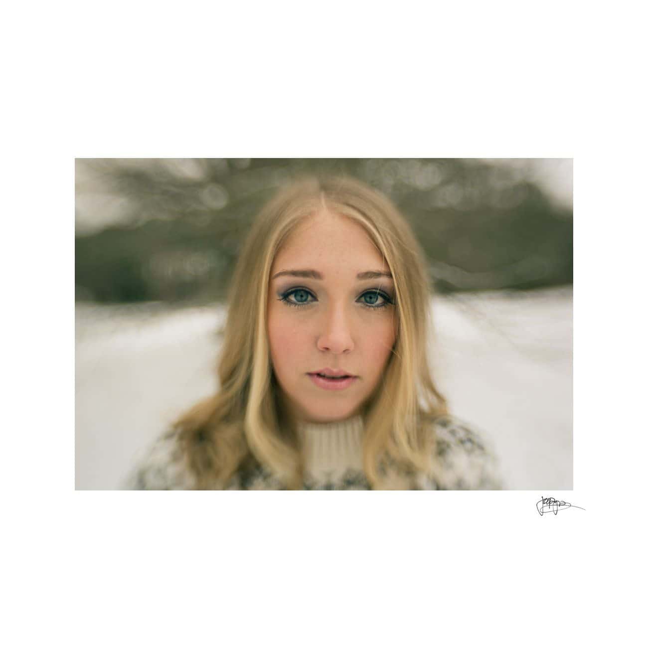 A woman with long blonde hair and blue eyes stands outdoors in a snowy landscape. She is wearing a patterned sweater and looks directly at the camera. The background is softly blurred, giving a dreamy effect to the setting.