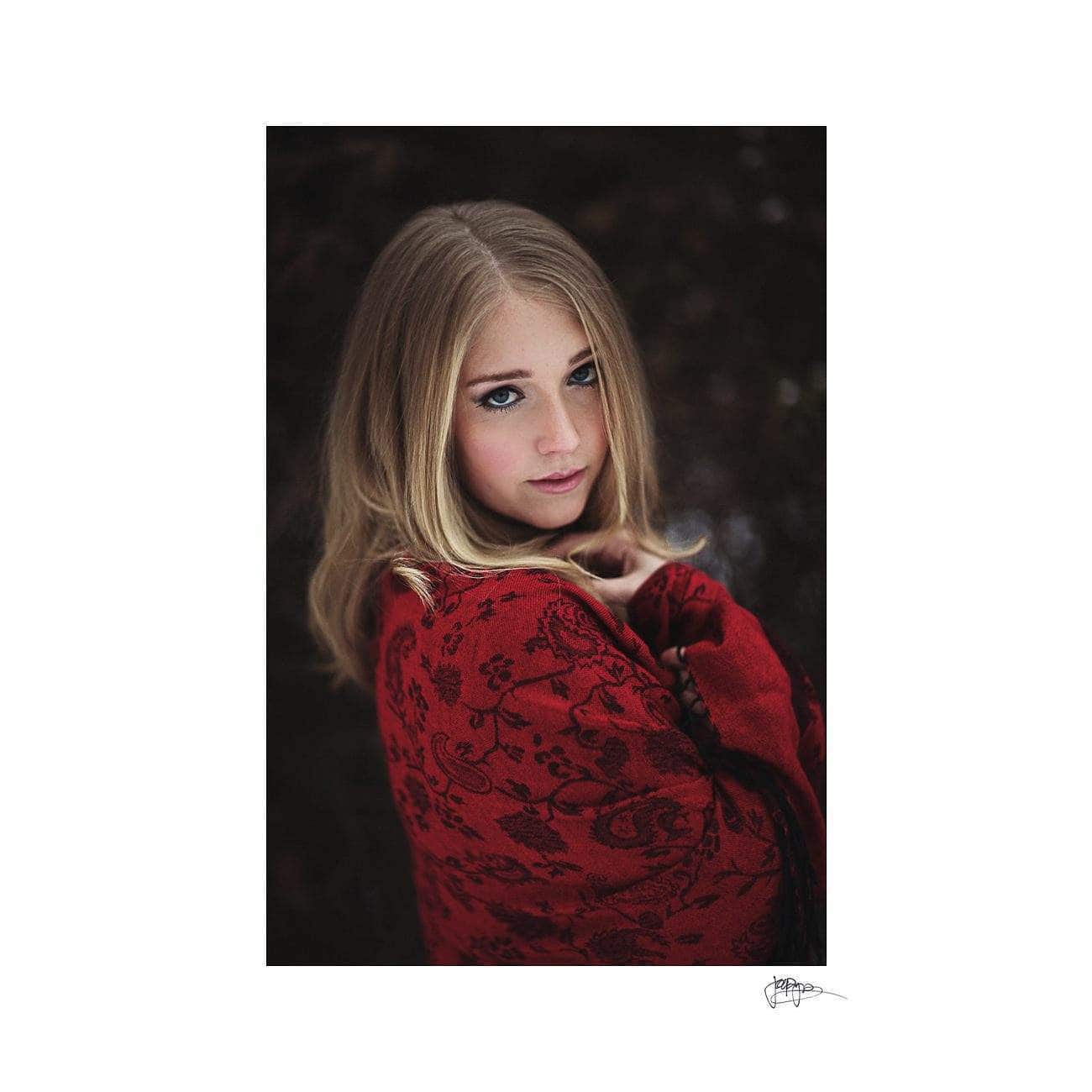 A woman with long blonde hair gazes into the camera. She is wrapped in a red, patterned shawl against a dark, blurred background.