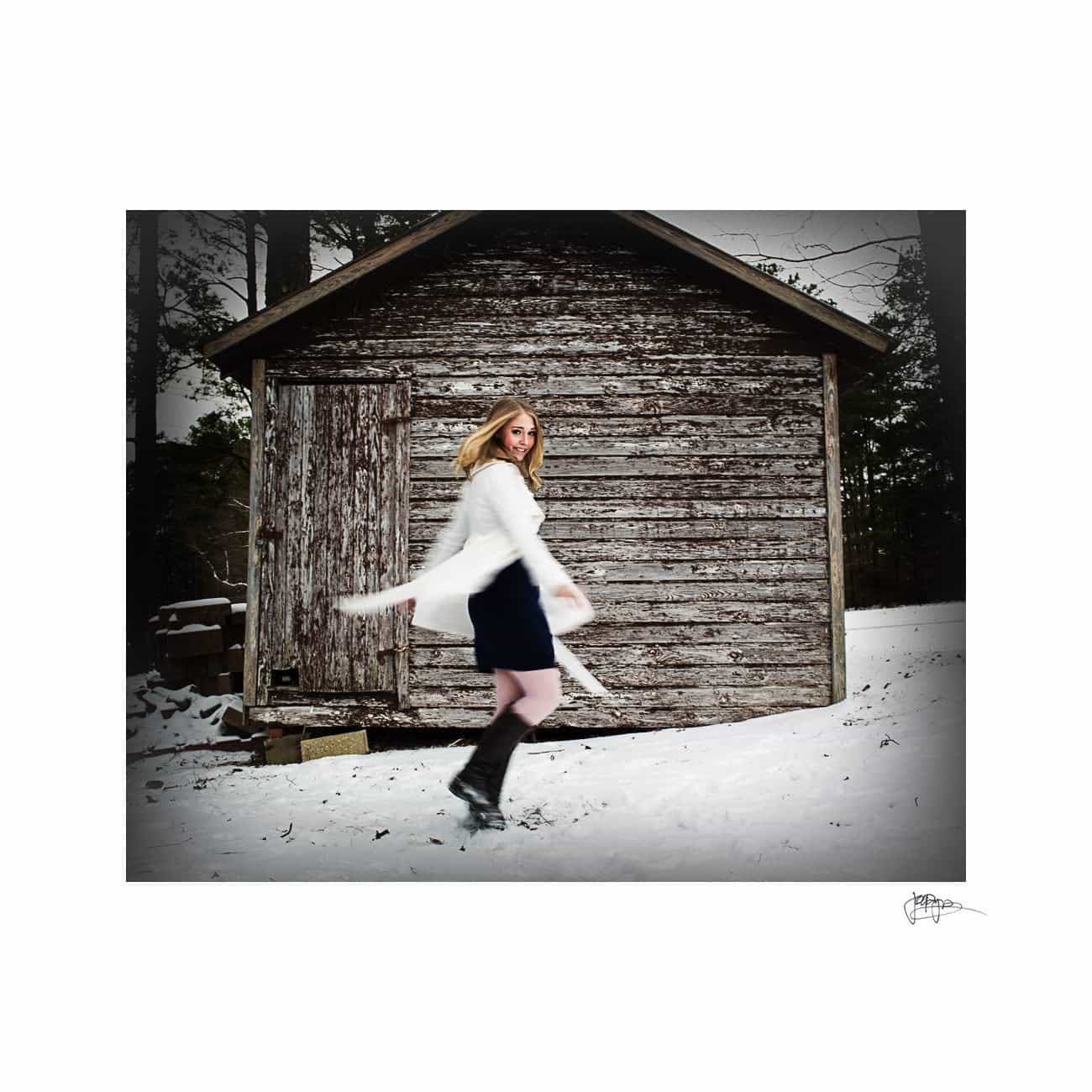 A person in a white coat and black skirt twirls joyfully in the snow in front of an old wooden shed. The scene is surrounded by trees under a cloudy sky. The snow adds a serene, wintry atmosphere.