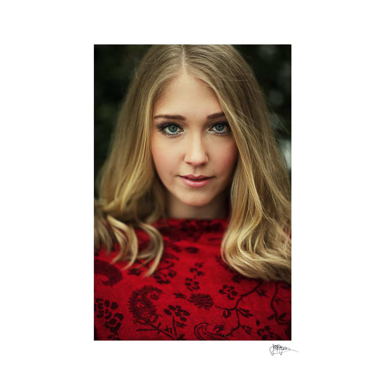 A person with long blonde hair is wearing a red patterned garment. They are looking directly at the camera with a neutral expression, and the background is softly blurred.
