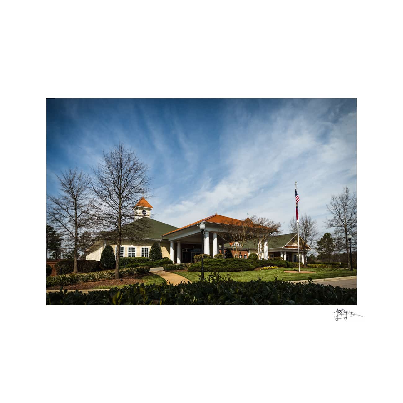 A picturesque building with a clock tower and a triangular roof, surrounded by trees and shrubs. An American flag stands prominently on a pole in the landscaped area under a partly cloudy blue sky.