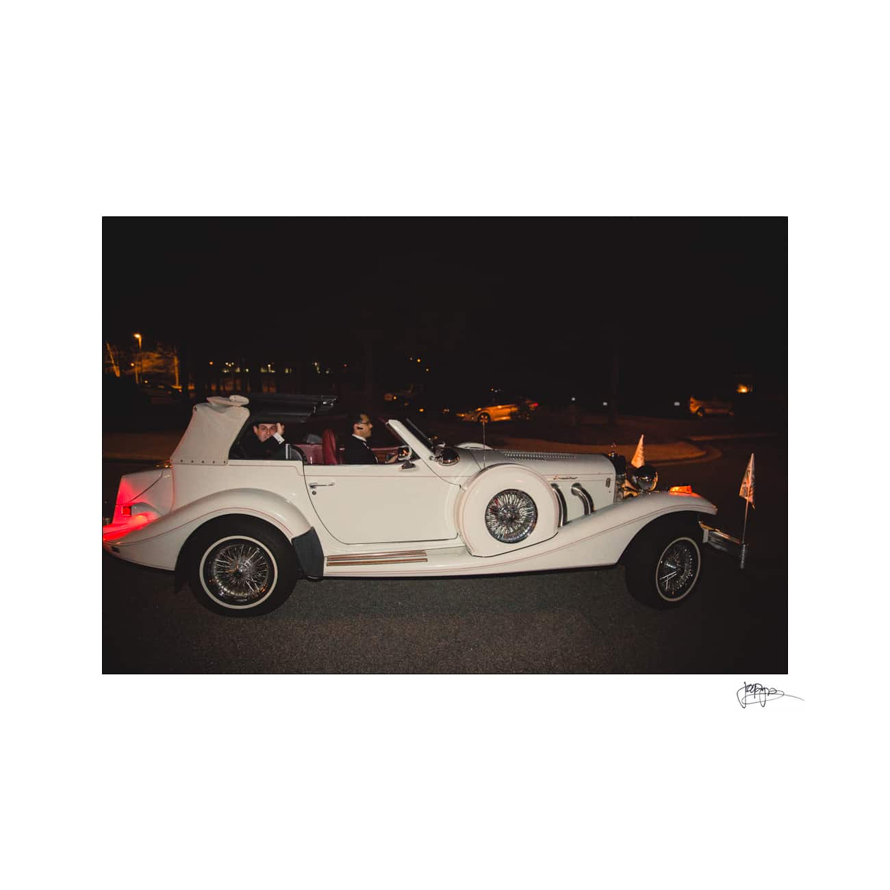 A vintage white convertible is parked at night on a road, with two people seated inside. The car features classic styling with wire wheels and side-mounted spare tires. The background is dimly lit, highlighting the cars elegance.