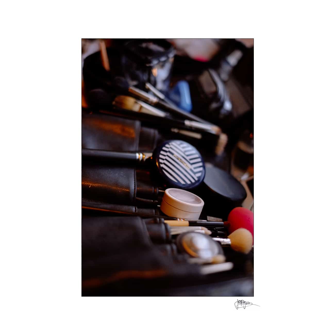 A close-up image of various makeup tools and products, including brushes, powders, and containers, arranged together. The focus is on the round compact with a striped design, surrounded by other beauty items.