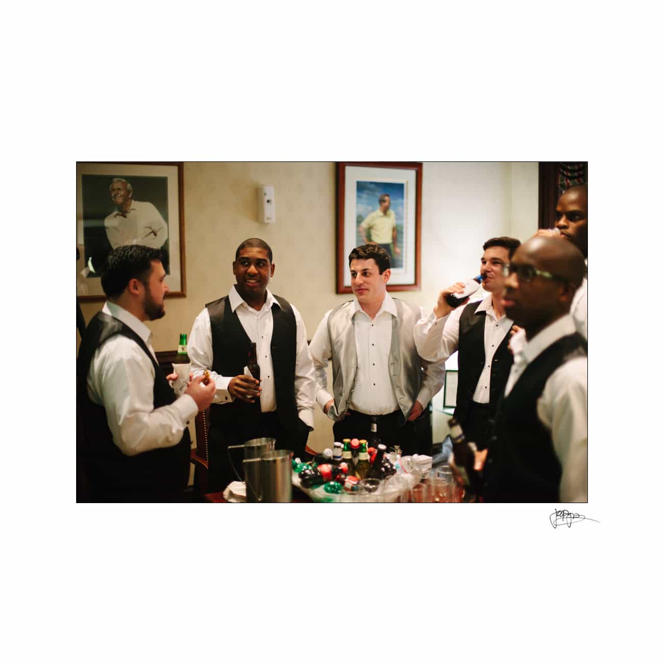 A group of six men in formal attire gather around a table with drinks and snacks. They appear to be socializing indoors. Two framed photos are on the wall behind them.