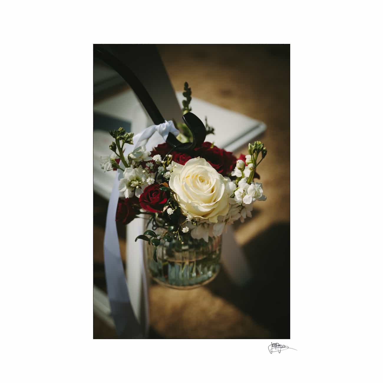 A small floral arrangement featuring a large white rose, surrounded by smaller red roses and white blossoms, is attached to a white chair with a white ribbon. The setup is outdoors on a sunny day.