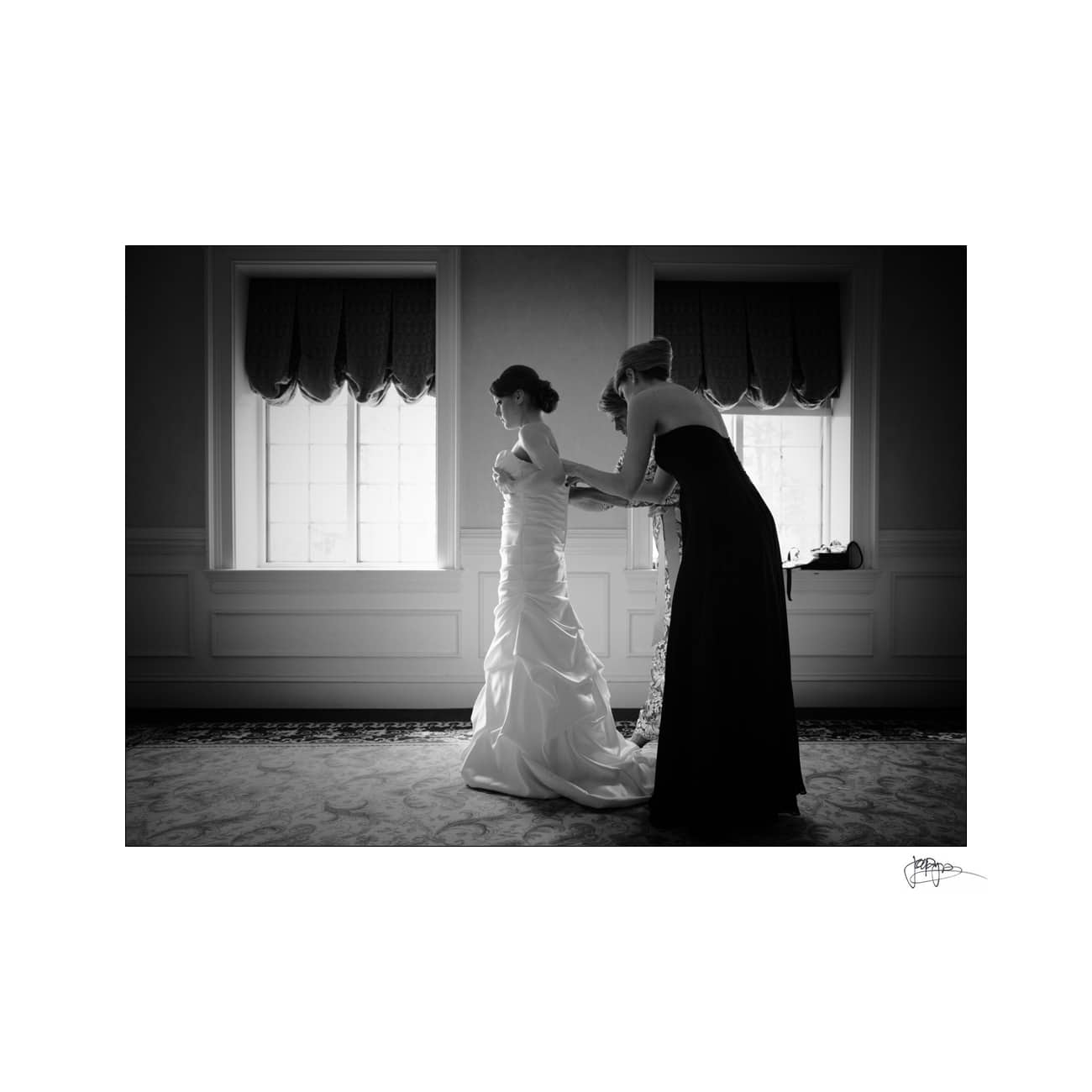 A black and white photo of a bride in a gown being helped with her dress by another woman in a long dress. They stand between two large windows with curtained valances, creating a serene and elegant atmosphere.