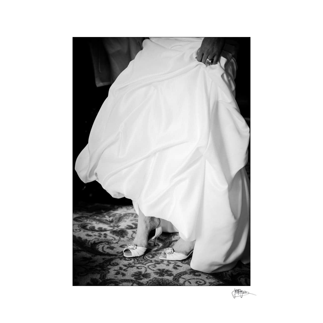 A black and white photo of a person wearing a flowing white dress, elegantly lifting it to reveal their feet. They are wearing white, open-toed shoes with a small heel, standing on a patterned carpet.