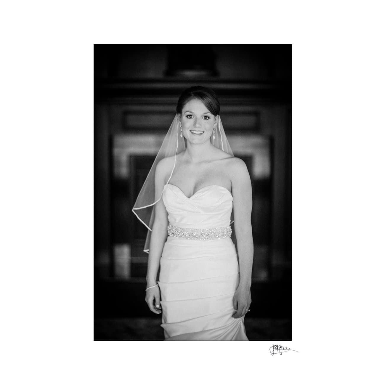 A black and white photo of a bride wearing a strapless gown with a beaded belt and a veil, smiling at the camera. The background is softly blurred, highlighting her.