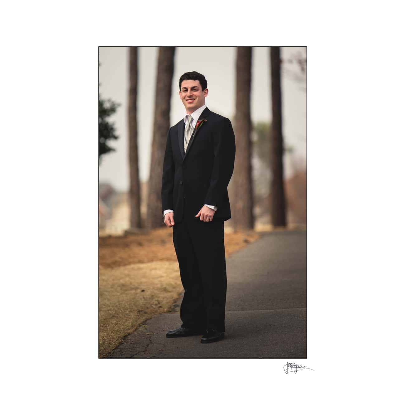 A young man in a black suit, white shirt, and striped tie stands on a path lined with trees. He smiles slightly, posing with his hands by his sides. The background is softly blurred, creating a serene outdoor setting.