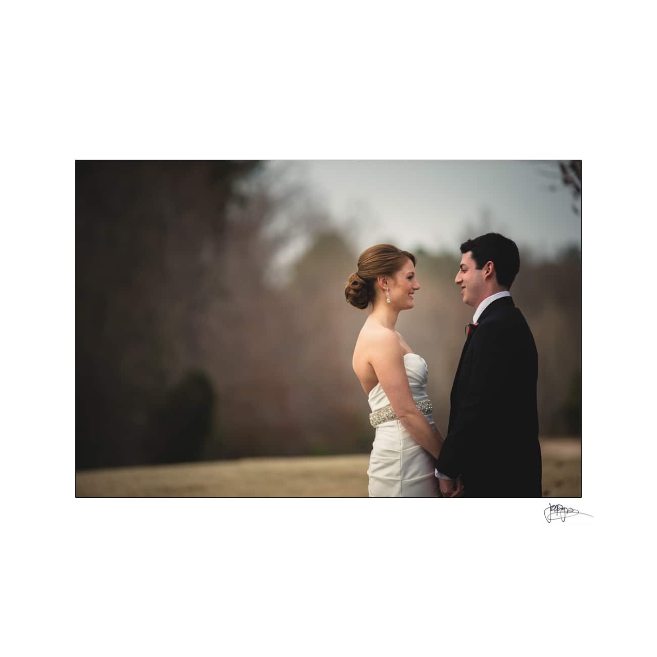 A bride and groom stand facing each other outdoors on a cloudy day. The bride wears a strapless white gown, and the groom is in a dark suit. They hold hands, surrounded by a blurred natural background.