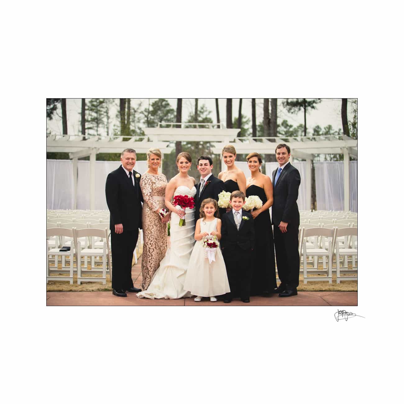 A group of nine people dressed in formal attire pose for a wedding photo. The bride, in a white gown, holds red flowers. The groom stands beside her. Two children are in front, smiling. The background features white chairs and greenery.