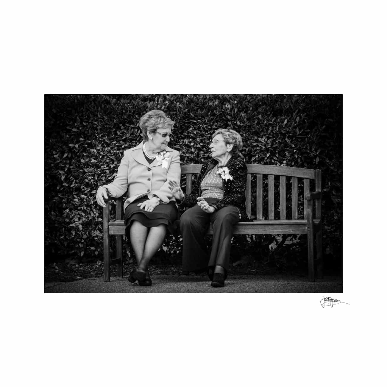 Two elderly women sitting on a wooden bench, engaged in conversation. They are both dressed in formal attire with floral corsages. The background features dense hedges. The image is in black and white.