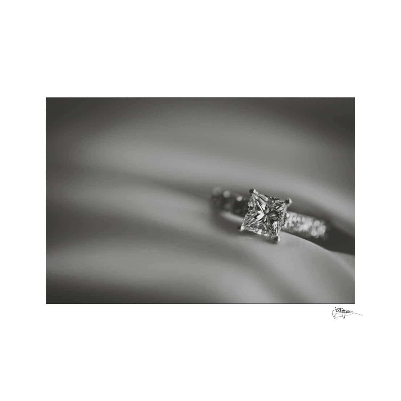 Close-up of a diamond engagement ring featuring a solitaire princess-cut diamond set on a smooth, textured surface. The image is in black and white, highlighting the clarity and detail of the diamond and band.