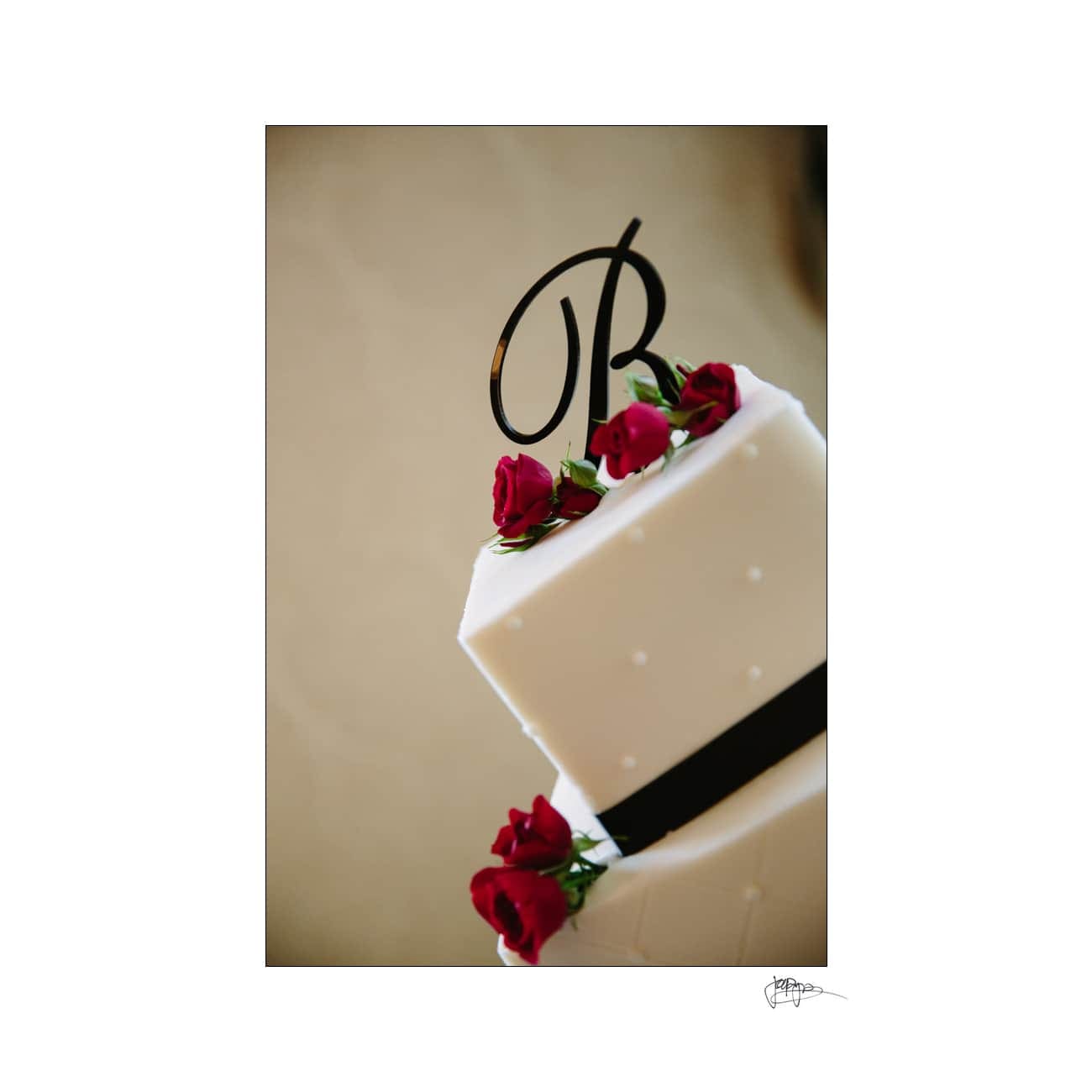 A white tiered cake decorated with small red roses and a black ribbon is topped with a black letter B.