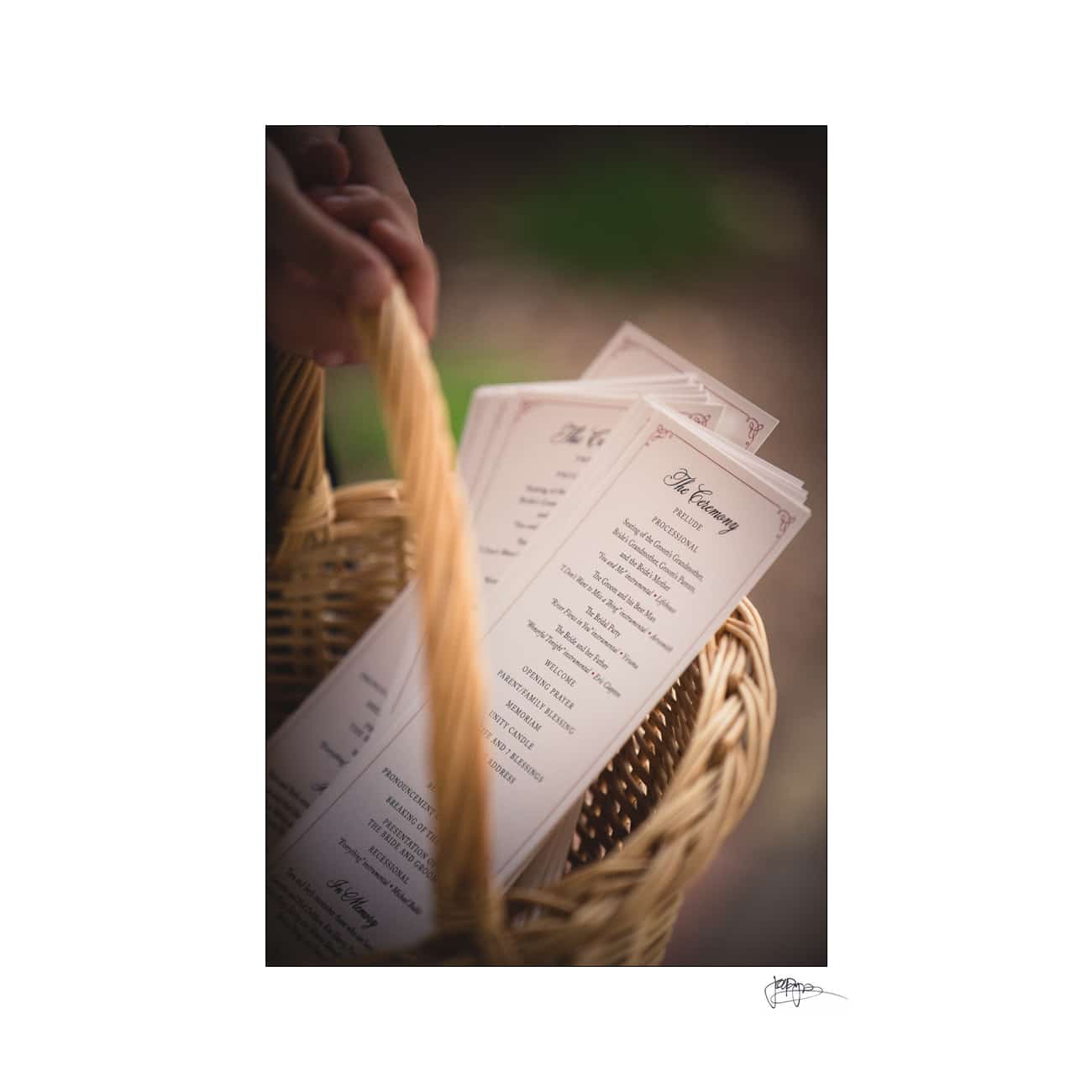 A hand holding a woven basket filled with wedding ceremony programs printed on cream-colored paper with elegant typography.