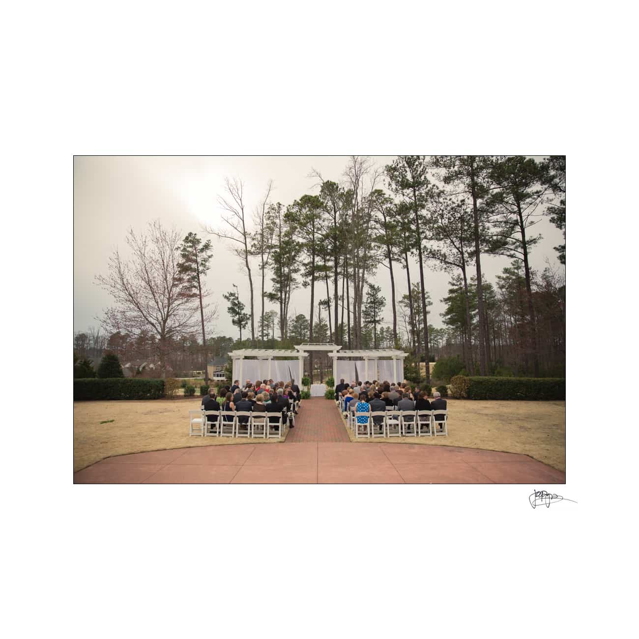 Outdoor wedding ceremony with guests seated on white chairs facing a floral-decorated altar. Tall trees and overcast sky in the background. The setting is a manicured garden with a pathway leading to the altar.