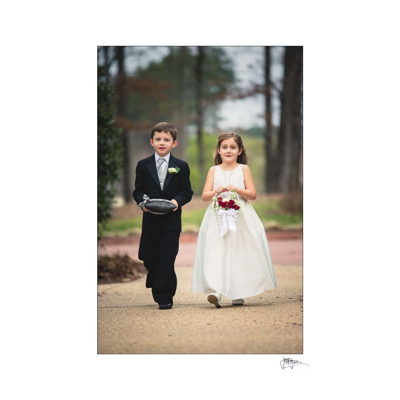 A young boy and girl are walking down an outdoor path. The boy is wearing a black suit and holding a pillow. The girl is in a white dress and holding a bouquet of red and white flowers. The background features trees and a pathway.