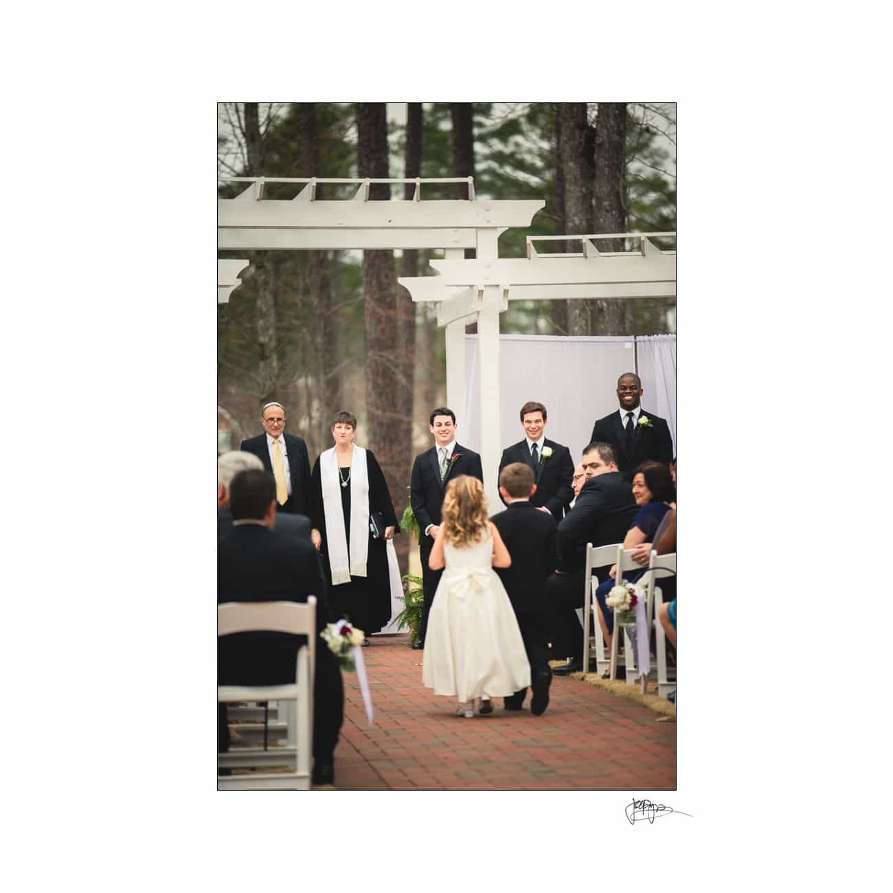 A flower girl with blonde hair walks down the aisle at an outdoor wedding, holding a basket. Guests sit along the aisle while the wedding officiant, the groom, and groomsmen stand at the altar under a white pergola.