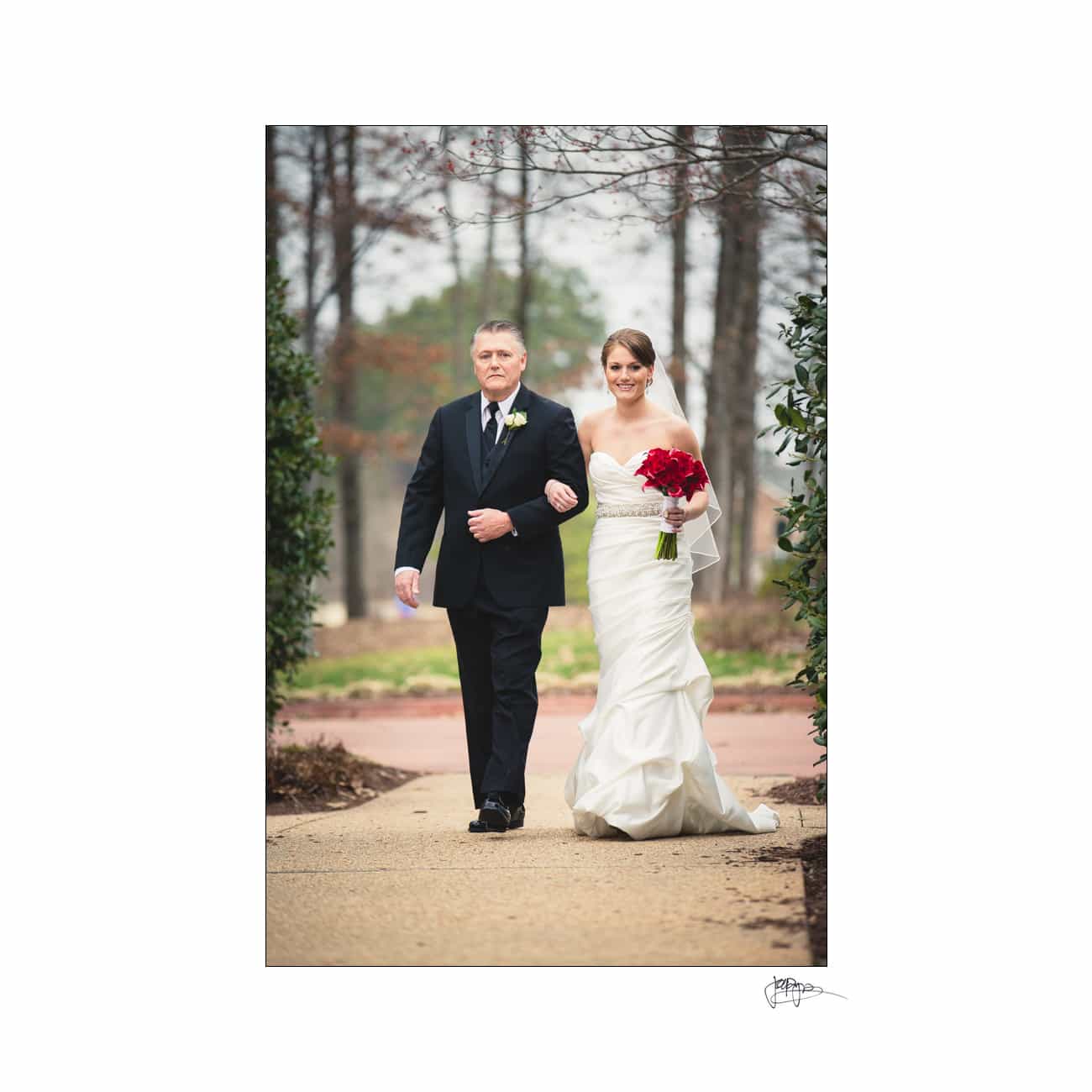 A bride in a white dress holds a red bouquet and walks arm-in-arm with an older man in a black suit. They are outdoors on a path, surrounded by trees. Both are smiling as they proceed.