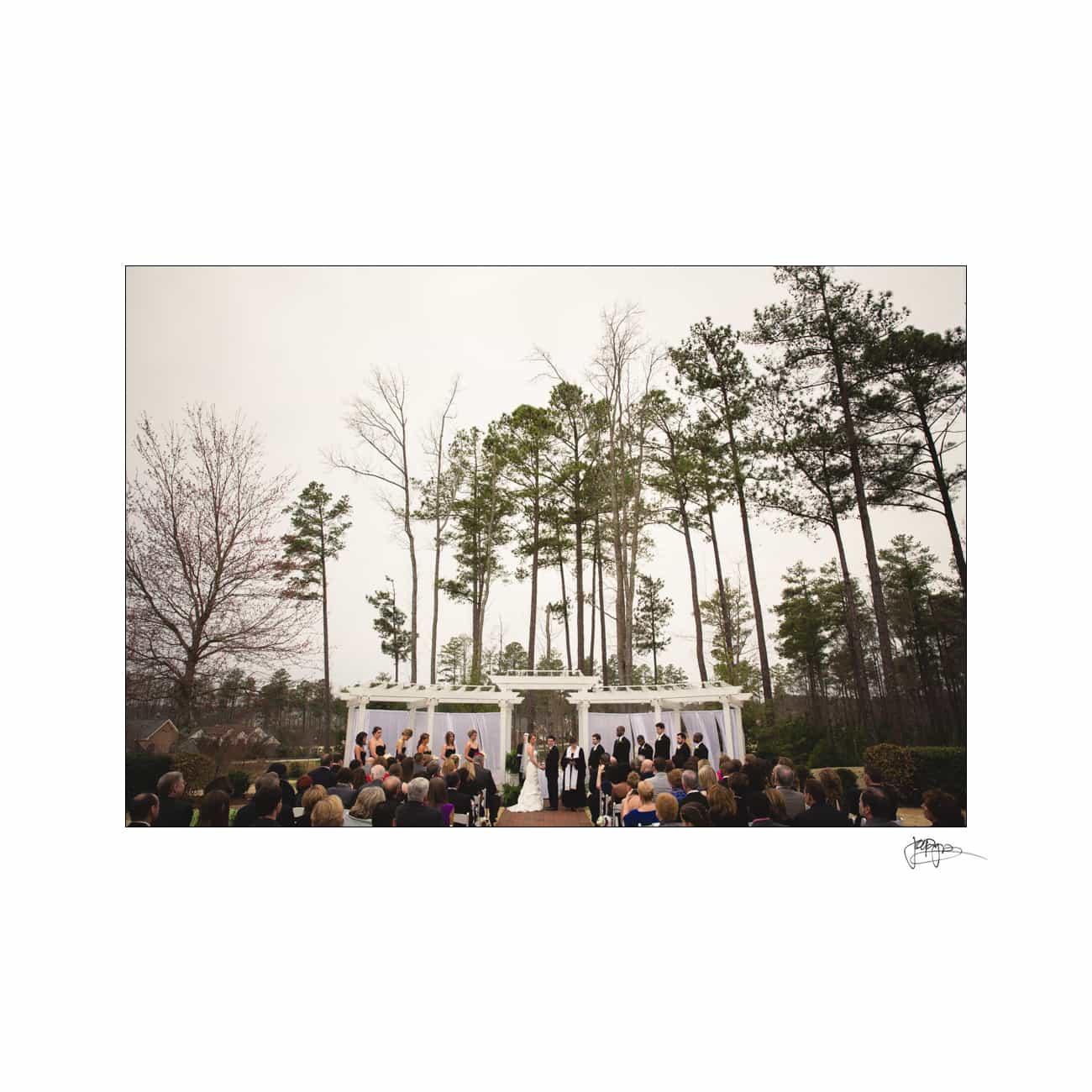 A wedding ceremony outdoors with a couple standing under a white gazebo. Bridesmaids and groomsmen are in formation on either side. Tall trees frame the scene, and guests are seated, watching the event. The sky is overcast.