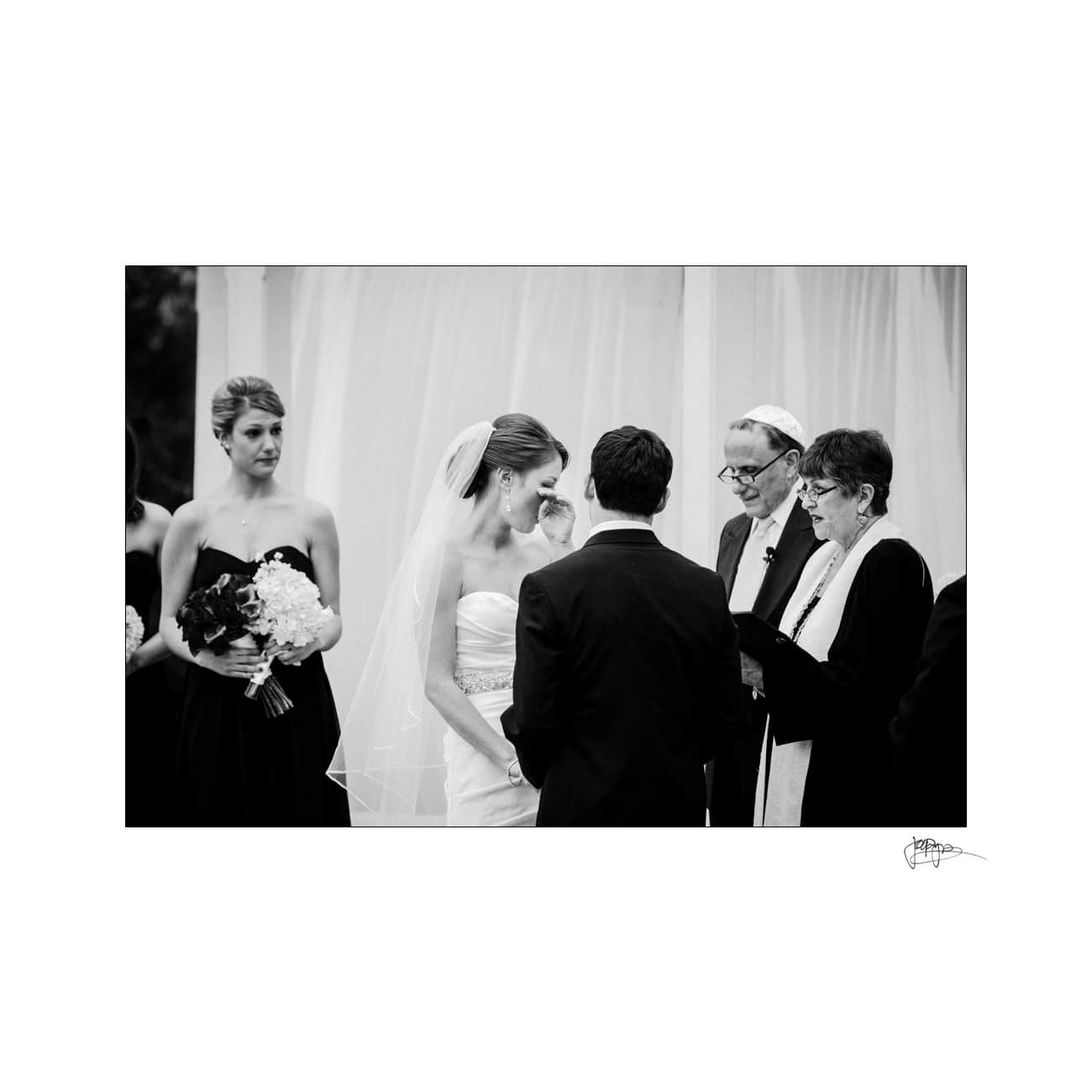 A black and white photo of a wedding ceremony, with the bride in a strapless gown and veil, the groom in a suit, and two officiants conducting the service. A bridesmaid stands to the side holding a bouquet.