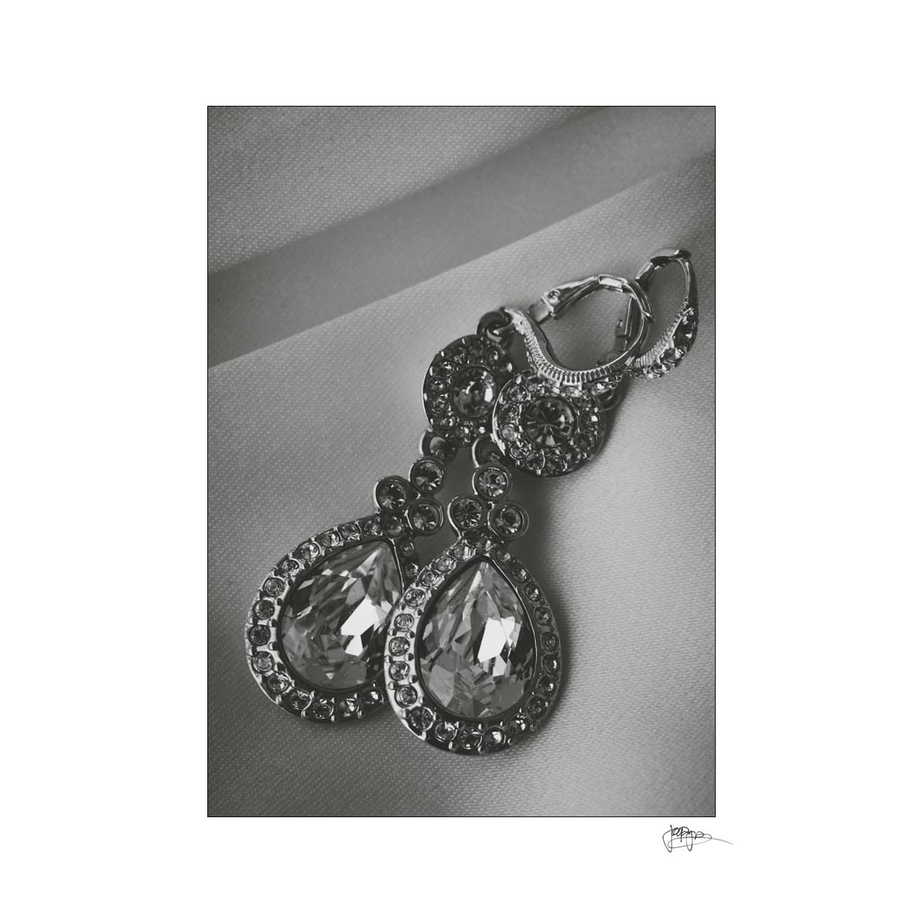 A pair of elegant teardrop earrings encrusted with sparkling gemstones, displayed on a smooth, light-colored fabric backdrop. The earrings feature intricate detailing and a shimmering finish.