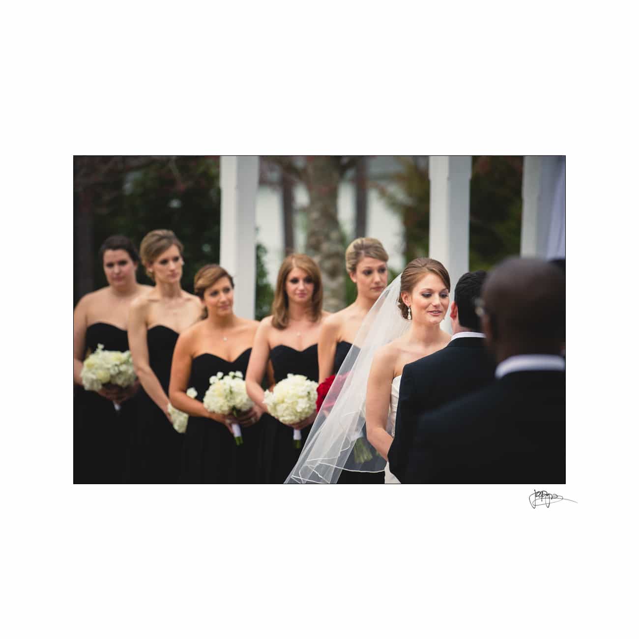 A bride and groom stand together during a wedding ceremony. The bride wears a white gown and veil, while the groom is in a suit. Bridesmaids stand nearby in black dresses, holding white bouquets.