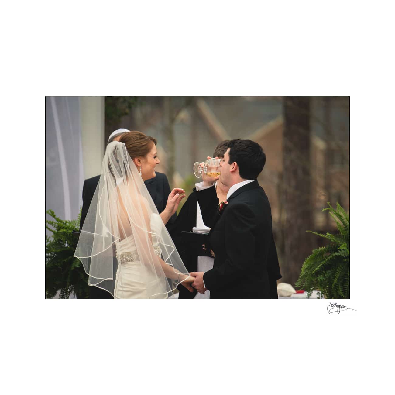 A bride and groom are standing together, holding hands during a wedding ceremony. The groom is drinking from a glass while a person officiating stands behind them holding a book. The bride is smiling. Ferns and drapery decorate the setting.
