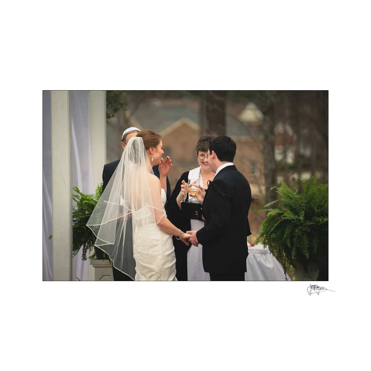 A bride and groom stand facing each other at an outdoor wedding ceremony. The bride, in a strapless gown and veil, holds the grooms hands. A minister and another individual stand beside them. Lush greenery and blurred background are visible.