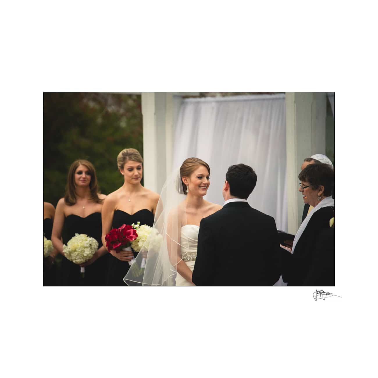 A bride and groom stand facing a rabbi during an outdoor wedding ceremony. The bride wears a veil and white dress, holding red flowers. Three bridesmaids in black dresses stand to the side, holding white bouquets. A white canopy is in the background.