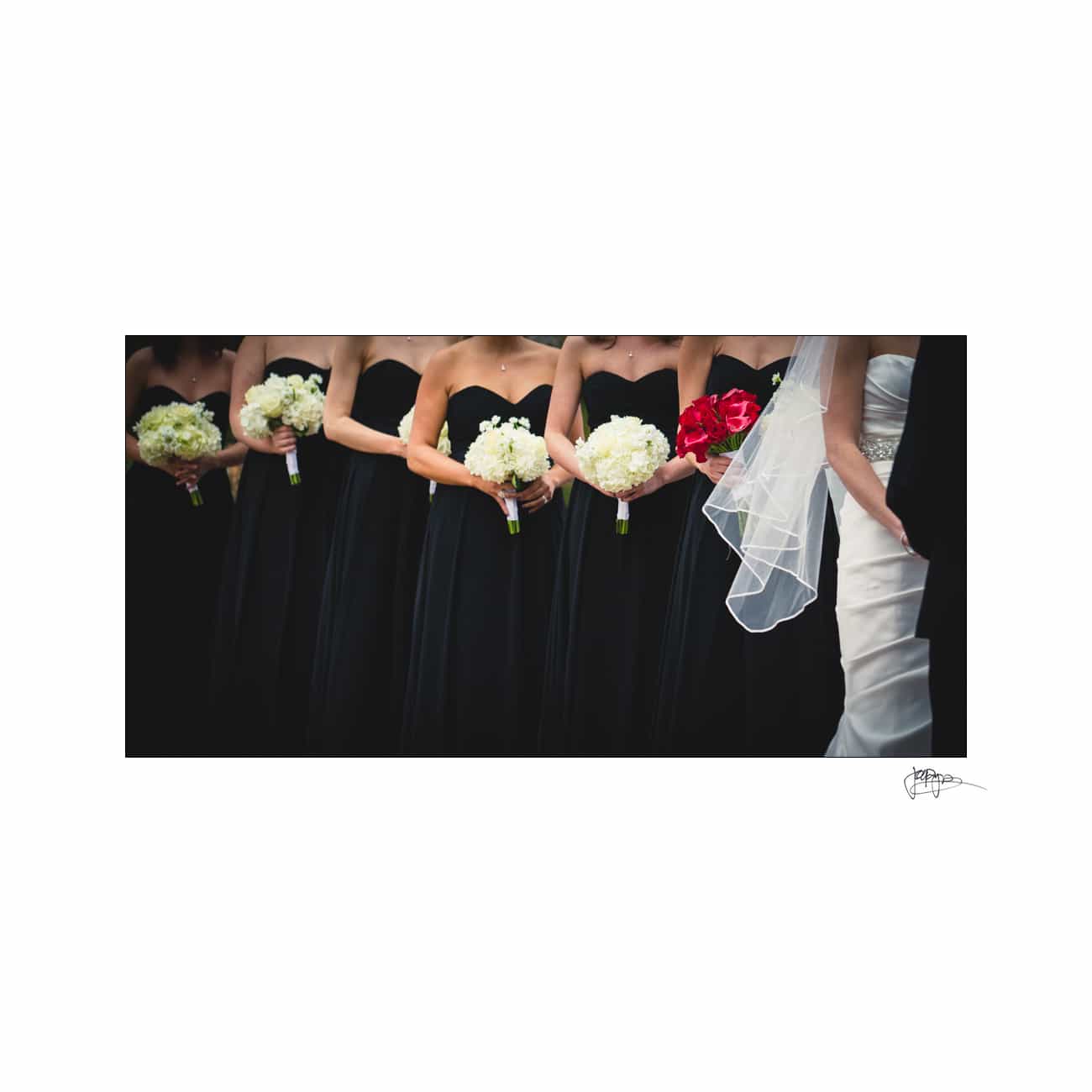 A line of bridesmaids in black dresses holds white bouquets, standing beside a bride in a white gown with a veil and red bouquet. Their faces are out of the frame, focusing on their outfits and flowers.