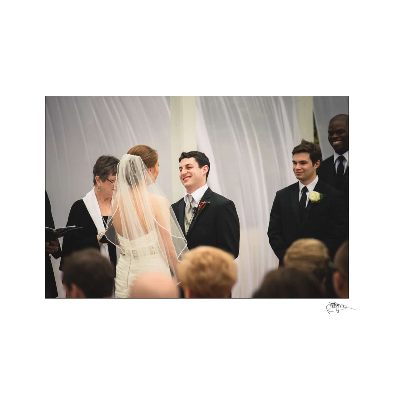 A bride and groom stand facing each other during a wedding ceremony. The bride wears a veil and white gown, while the groom is in a suit. Guests are seated in the foreground, and groomsmen stand nearby, smiling.