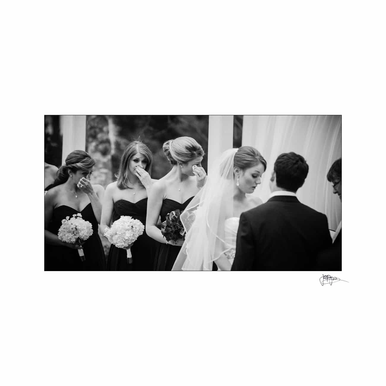 A black and white image of a wedding ceremony. The bride and groom stand facing each other, while bridesmaids in matching dark dresses hold bouquets and wipe their tears, suggesting an emotional moment.