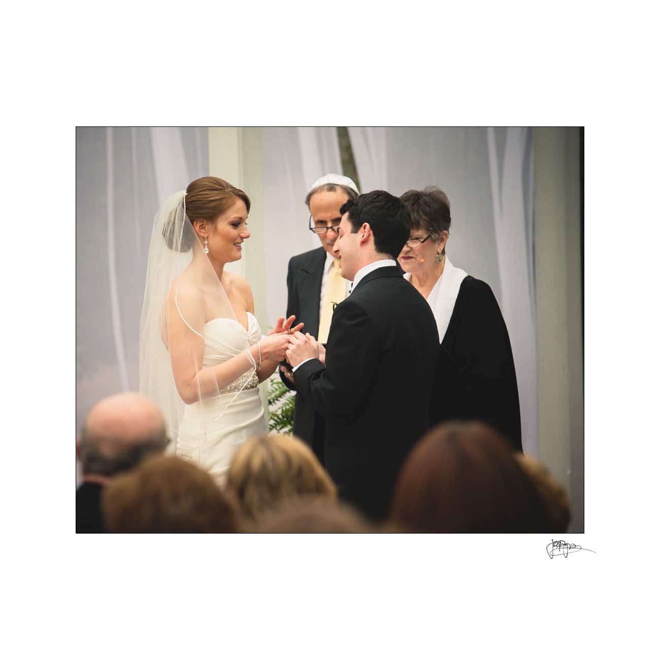 A bride and groom exchange rings during their wedding ceremony. The bride is wearing a white gown and veil, while the groom is in a black suit. Two officiants stand behind them in a decorated venue.