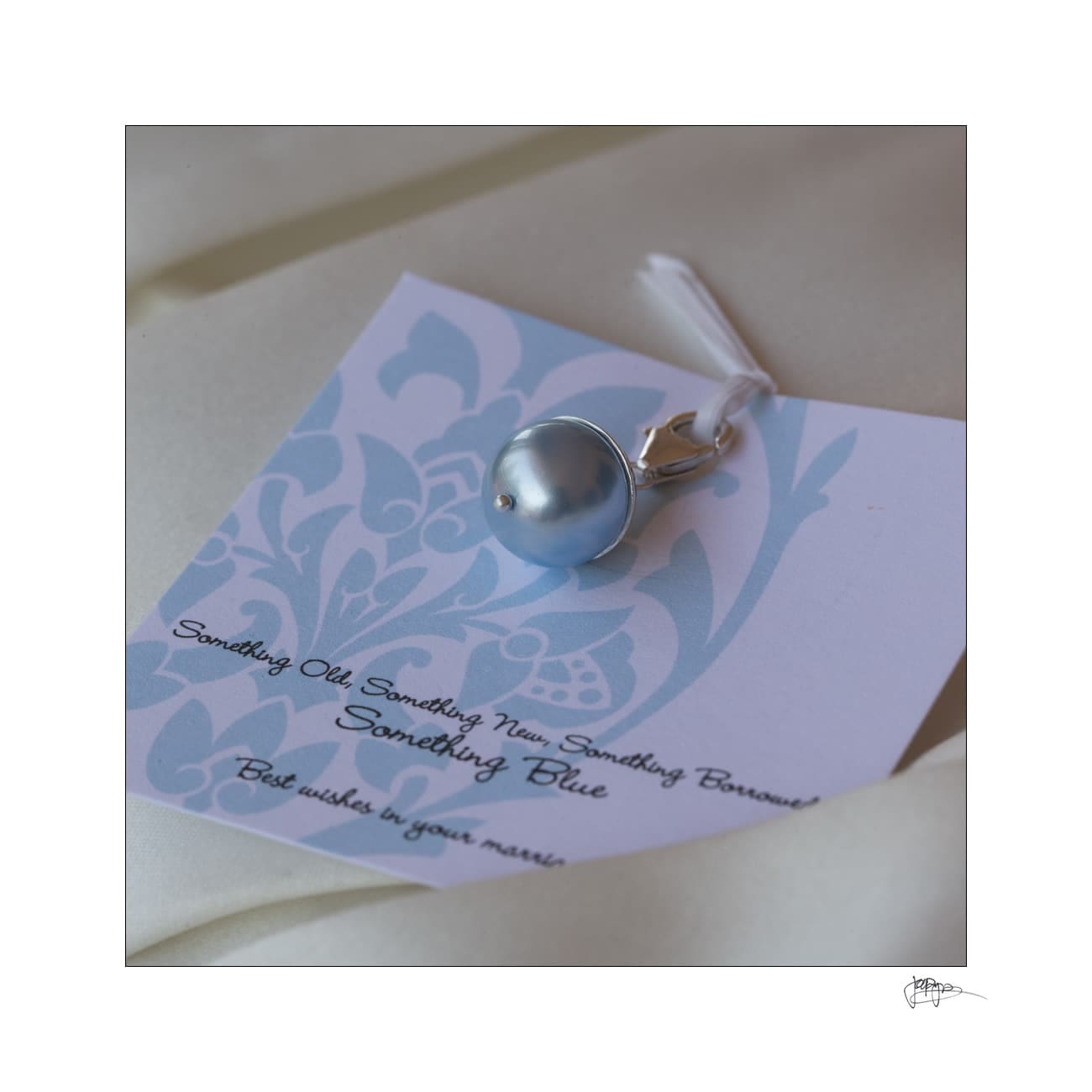 A blue pearl pendant attached to a card with a floral pattern. The card reads: Something Old, Something New, Something Borrowed, Something Blue. Best wishes on your marriage. The background is a soft, light fabric.