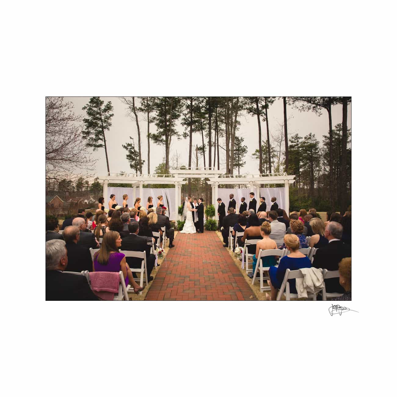 A wedding ceremony takes place outdoors on a brick pathway lined with guests. The couple stands under a white pergola, surrounded by trees and greenery. Bridesmaids in black dresses and groomsmen in suits are positioned on either side.