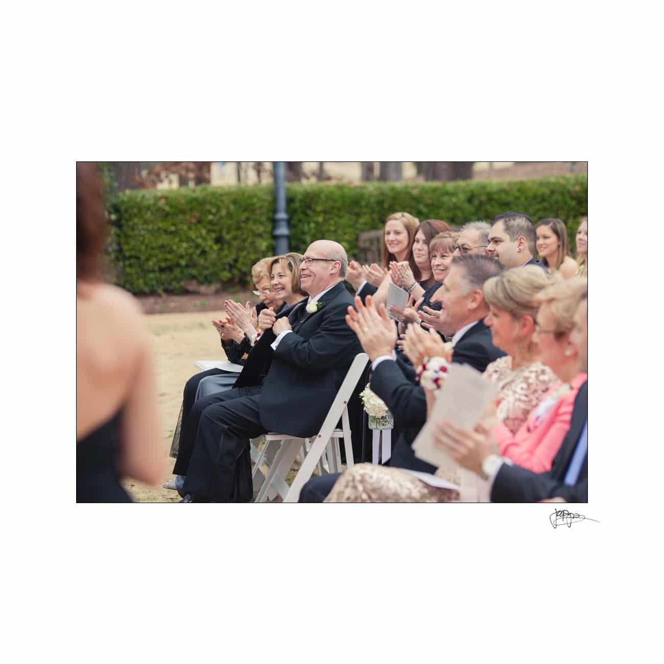 A group of people sits outdoors clapping and smiling. They are dressed in formal attire, suggesting a celebratory event, like a wedding or ceremony. Greenery in the background and a cloudy sky sets the scene.