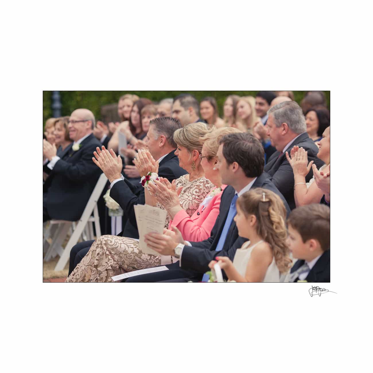 A diverse group of people at an outdoor event, seated in rows, clapping and smiling. Attendees include men, women, and children in formal attire. Some hold programs, and the background shows additional guests and greenery.