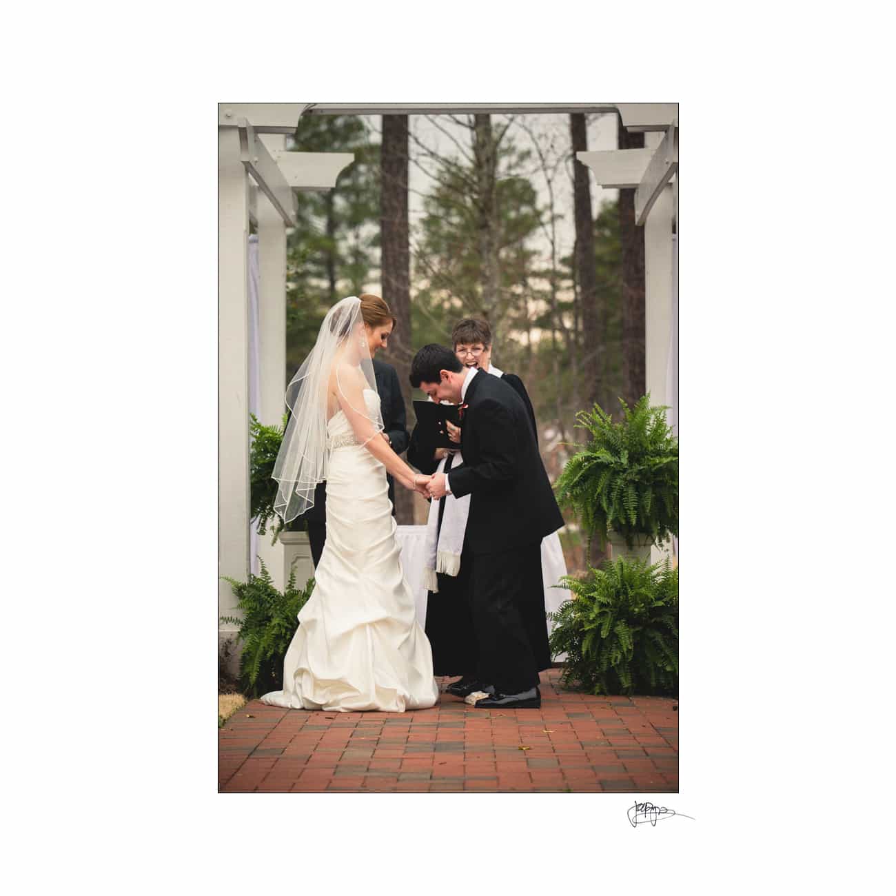 A bride and groom hold hands and exchange vows under a white pergola, surrounded by greenery. The groom leans down toward the bride. A person stands behind them, officiating the ceremony. Its an outdoor wedding with lush ferns decorating the scene.