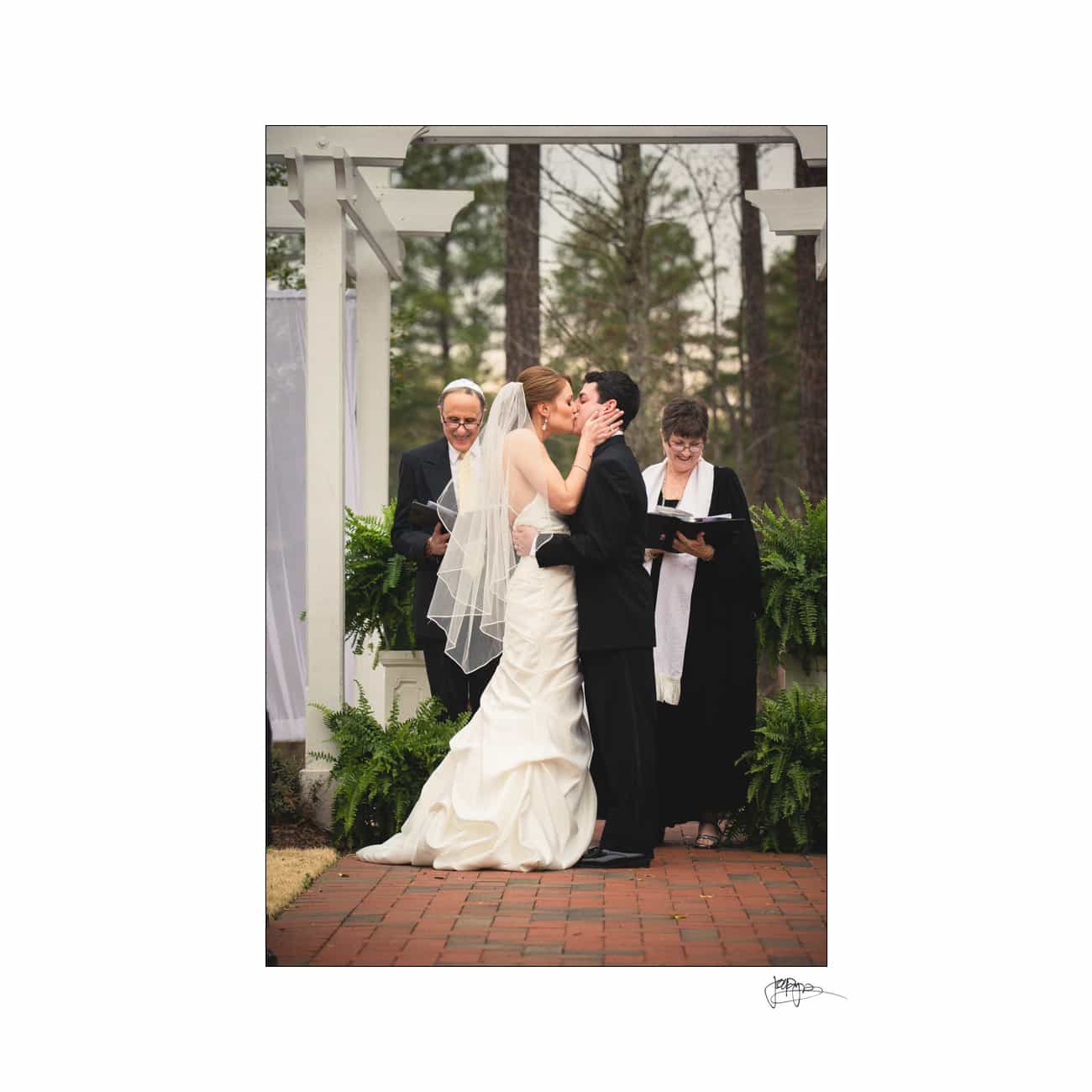 A bride and groom share a kiss at their wedding ceremony in an outdoor setting. They are under a white arbor, surrounded by greenery. Two officiants in black robes stand behind them, smiling.