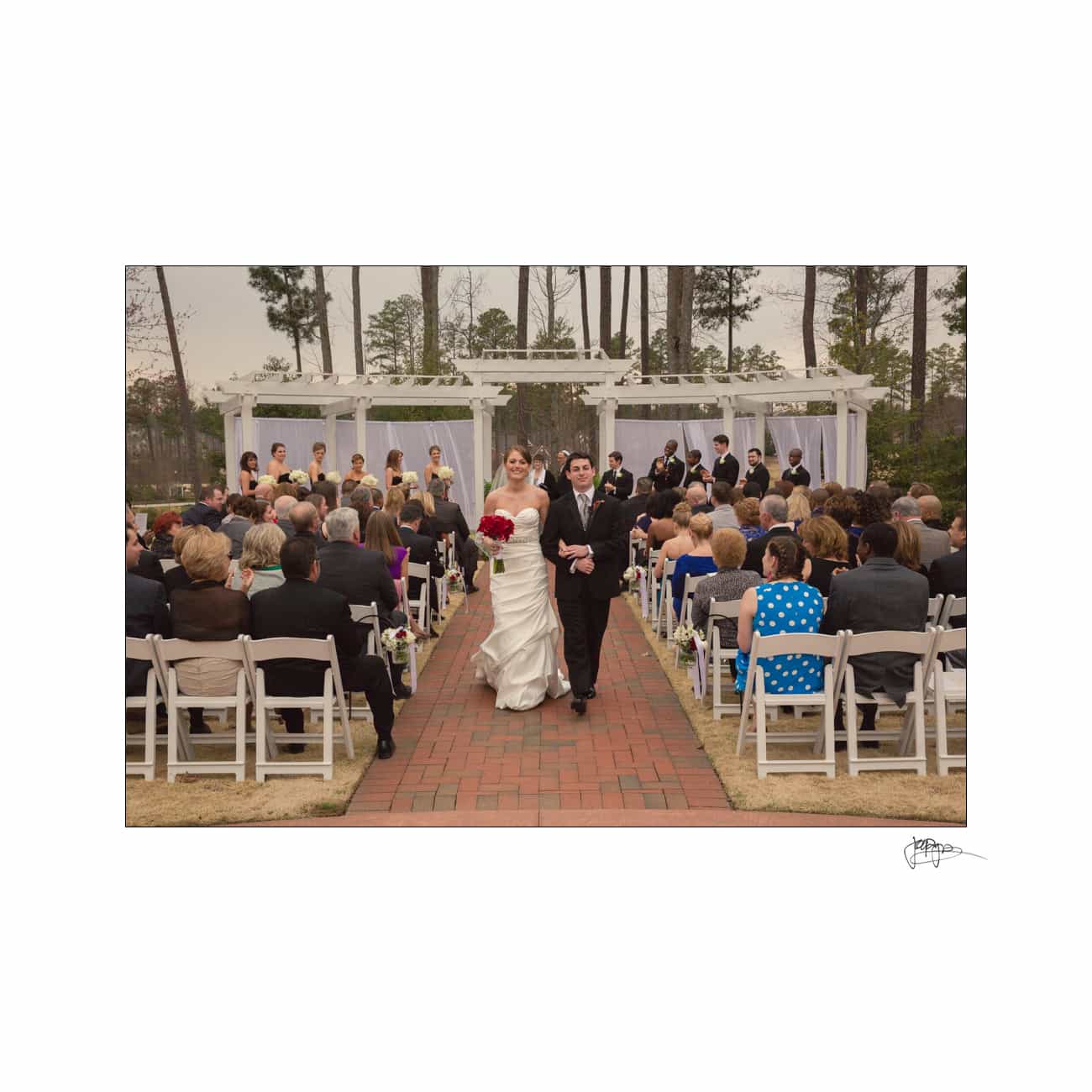 A bride and groom walk down the aisle outdoors under a white pergola, surrounded by guests seated in white chairs. The bride holds a red bouquet, and the wedding party stands in the background. The setting is lush and wooded.