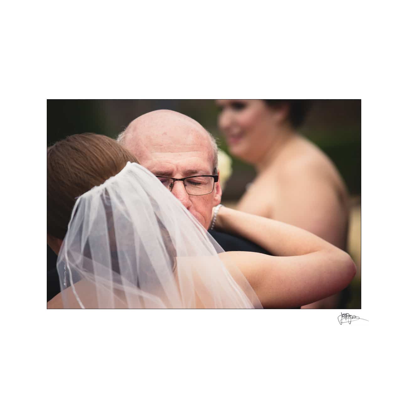 A bride, seen from behind wearing a veil, embraces an older man with glasses, possibly her father, in an emotional moment. A blurred figure wearing formal attire is visible in the background.