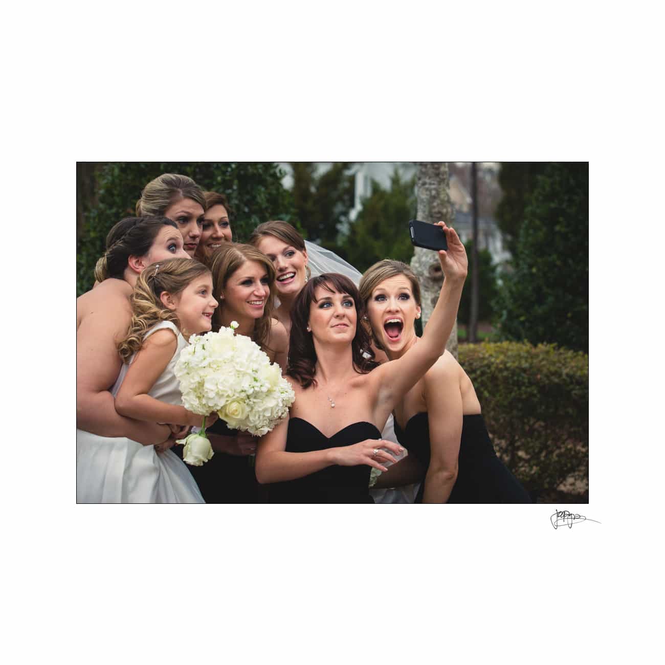 A group of smiling women, some in black dresses and others in white, gather closely for a selfie. One woman holds a bouquet of flowers. They are outdoors with greenery in the background.