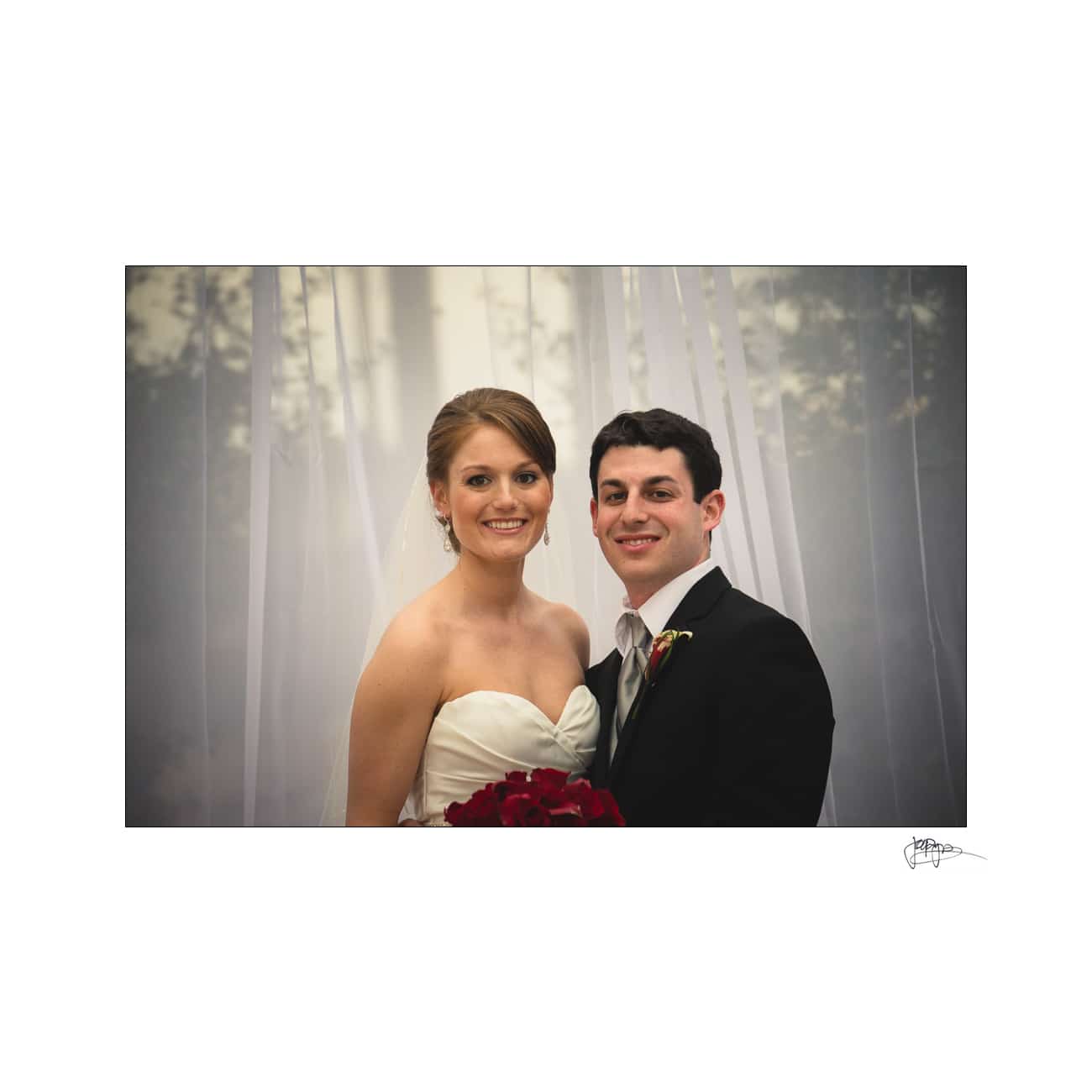 A bride in a strapless white gown and a groom in a black suit stand close together, smiling. The bride holds a bouquet of red flowers. The background is softly blurred, and there is an elegant, sheer curtain behind them.