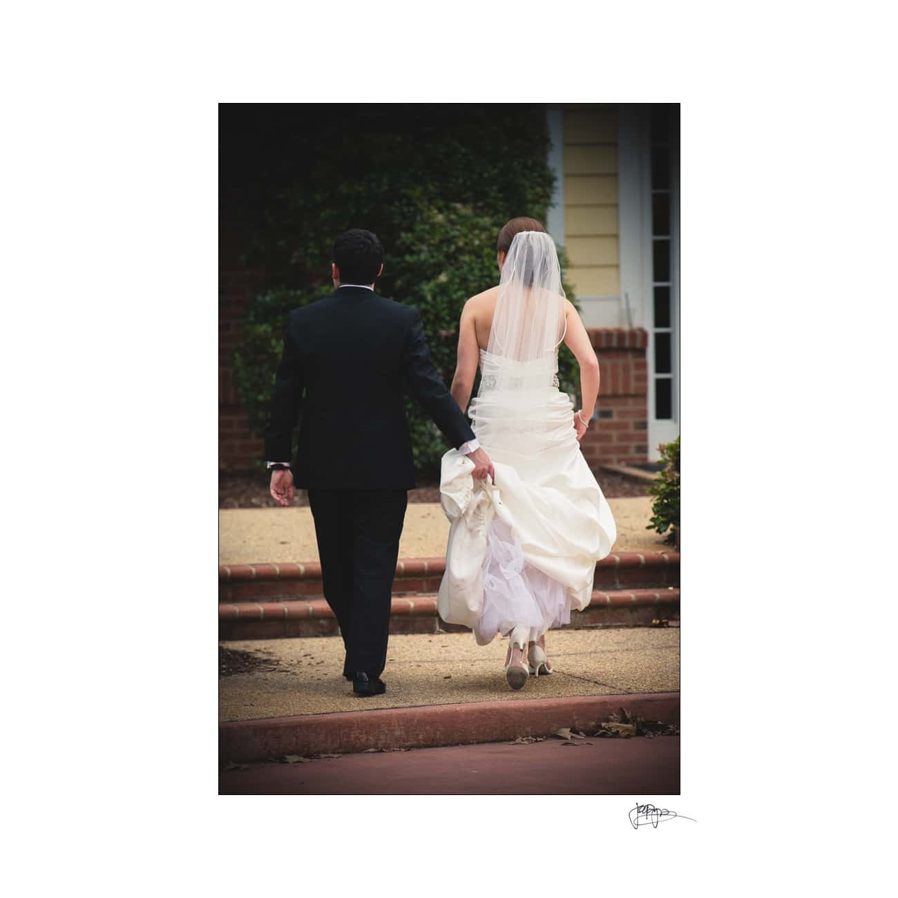 A bride and groom walk hand in hand away from the camera. The bride wears a white gown and veil, holding her dress with one hand. The groom is in a dark suit. Theyre walking on a paved path in an outdoor setting with brick and greenery in the background.