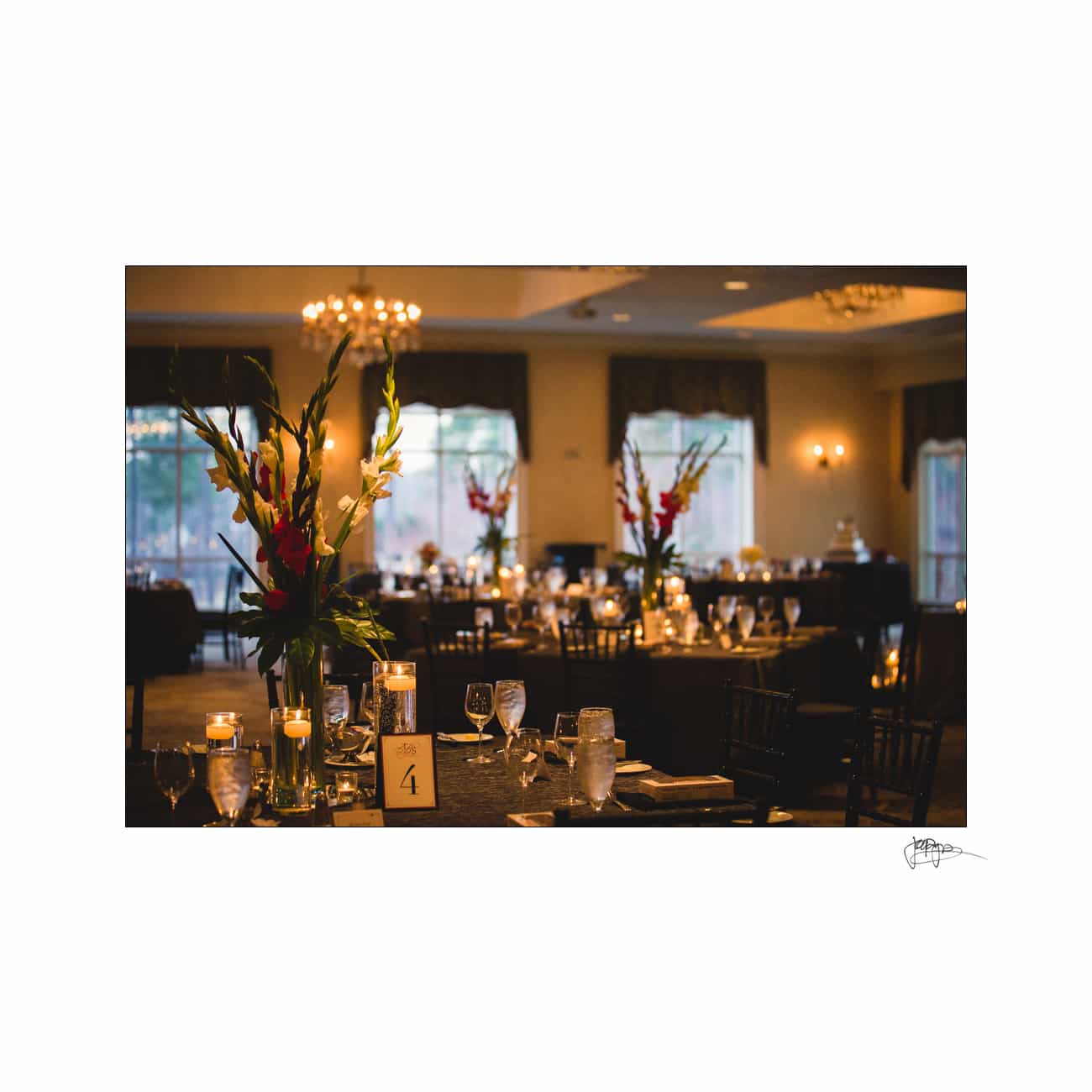 A dimly lit, elegant dining room set up for an event. Tables are adorned with candles, glasses, and floral centerpieces. Large windows in the background allow soft natural light, while chandeliers add warmth to the ambiance.