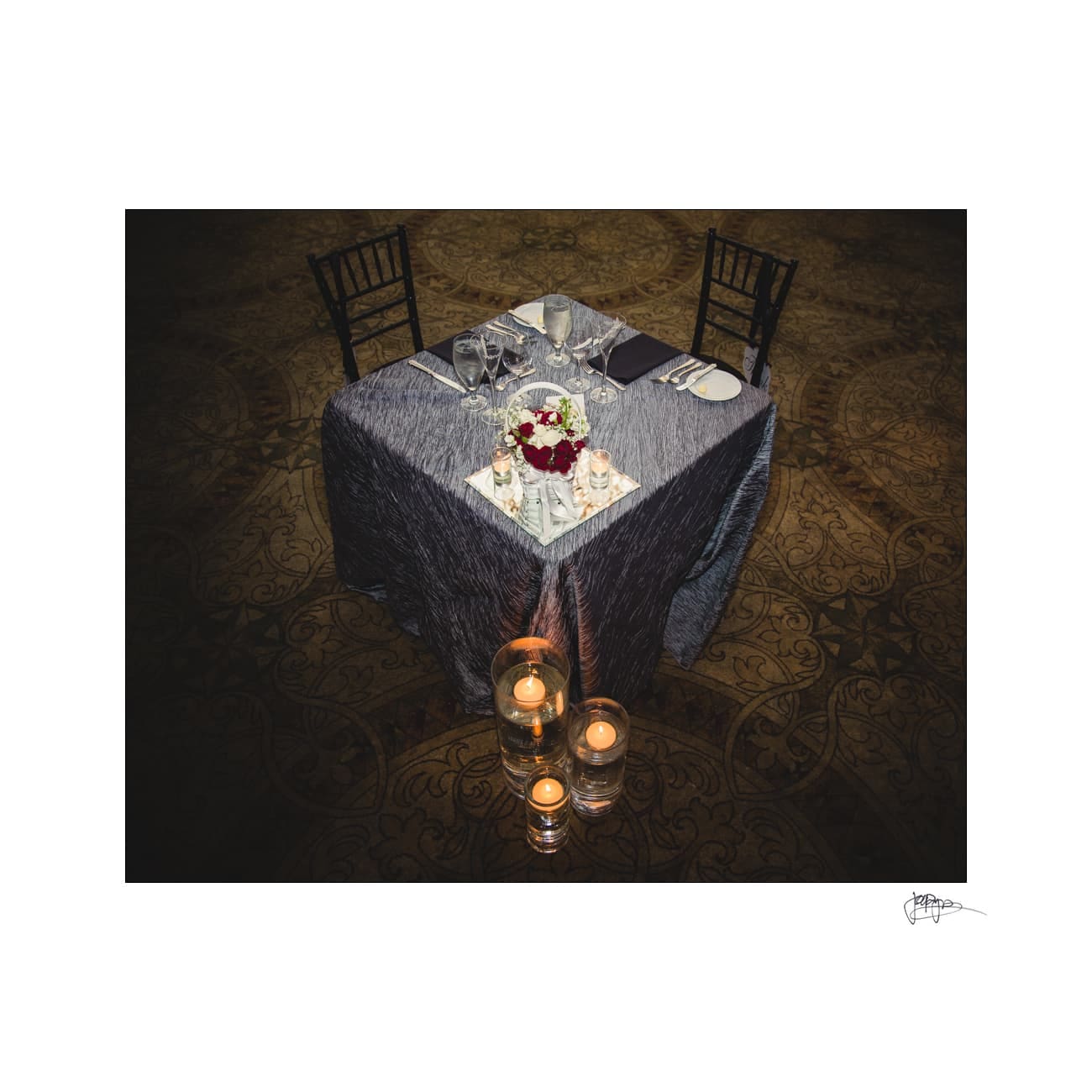 A small, elegantly set table for two with a gray tablecloth, adorned with a red and white floral centerpiece. Glassware and plates are neatly arranged, surrounded by three lit candles on the floor, creating a romantic ambiance.