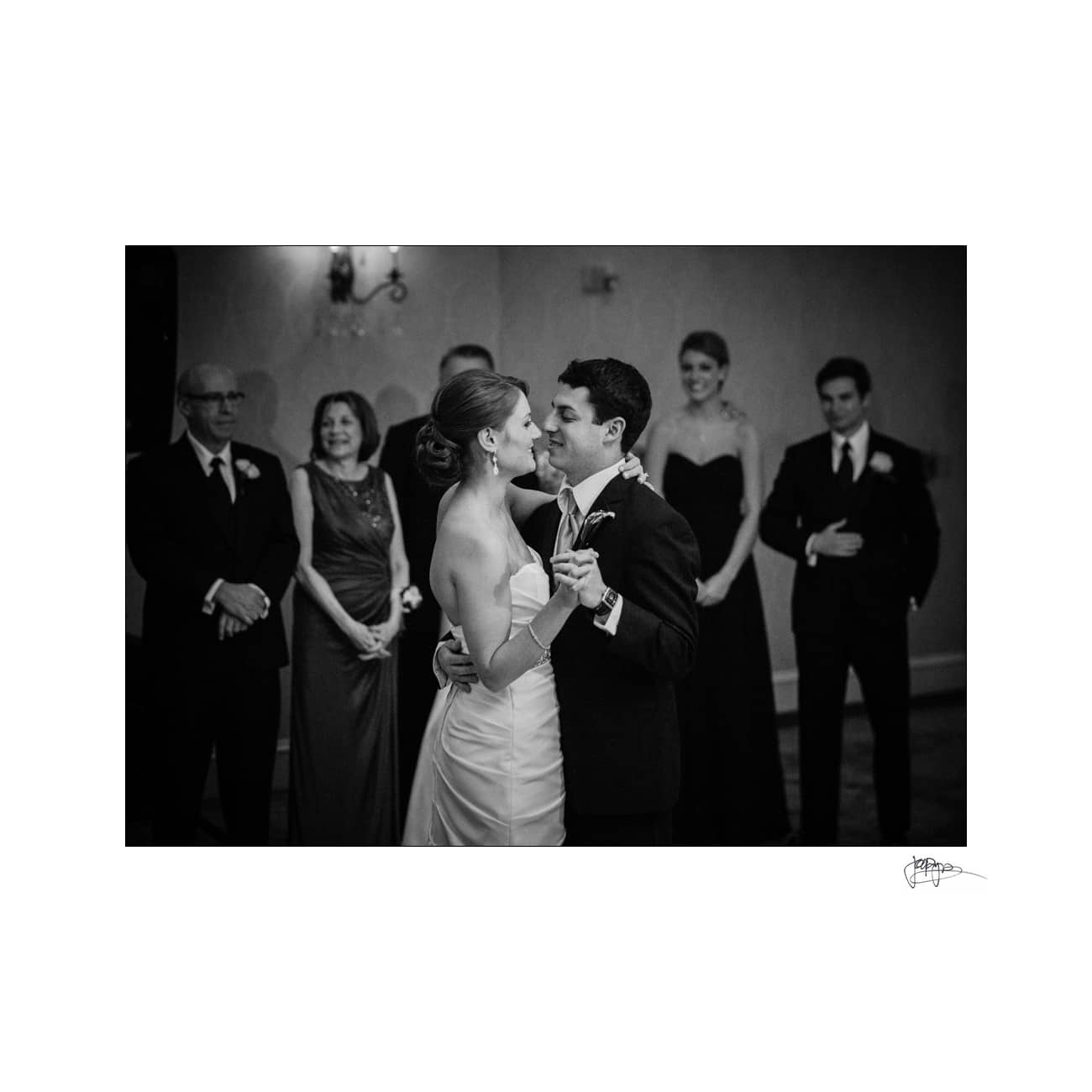 A bride and groom share their first dance at a wedding reception, surrounded by smiling family and friends. The bride is wearing a strapless gown, and the groom is in a suit and tie, looking at each other lovingly.