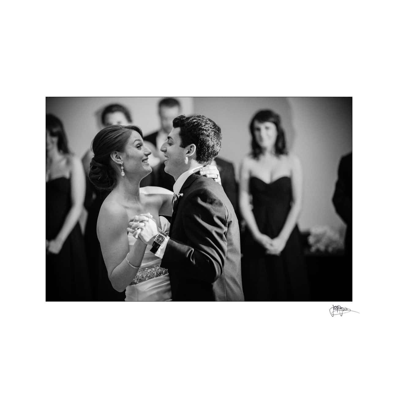 A bride and groom share their first dance, smiling as they look into each others eyes. The bride wears a strapless gown, and the groom is in a suit. Bridesmaids in dark dresses stand in the background, blurred. Black and white photo.