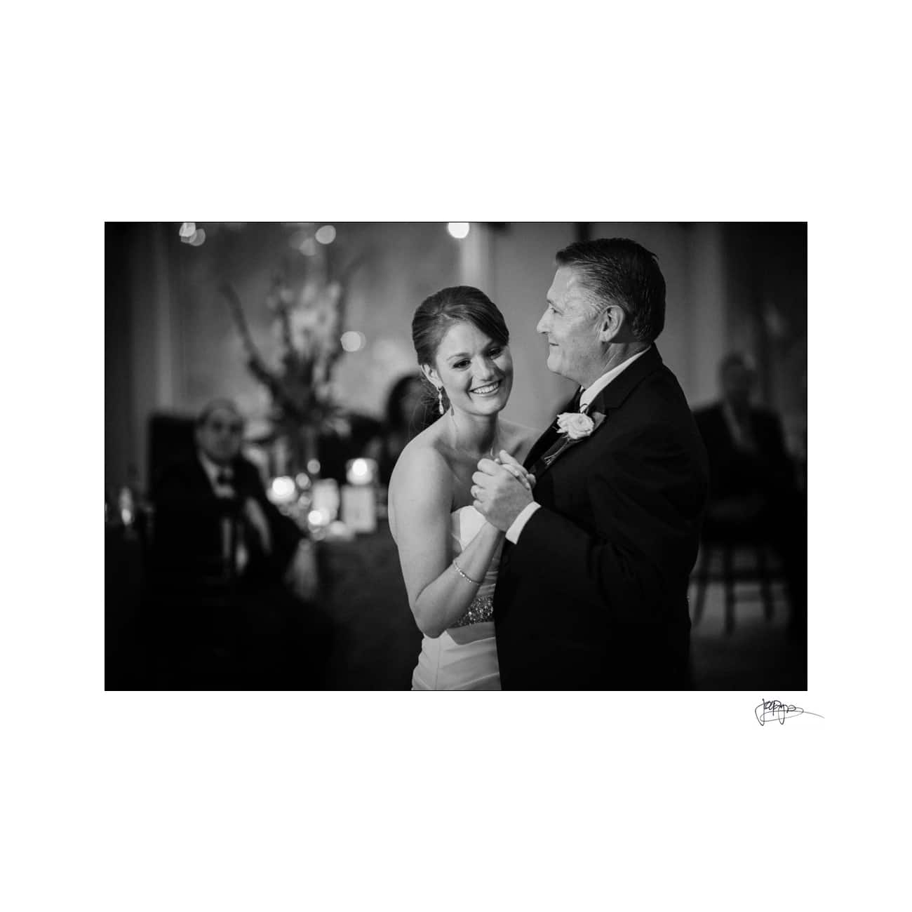A smiling bride in a white dress dances with an older man in a suit and tie at a wedding reception. The background is softly lit, with guests seated at tables.